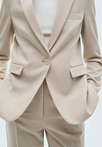 Veste beige à fermeture à un bouton, avec deux poches avant et un revers cranté ; associée à un pantalon beige assorti. Texture de tissu lisse.