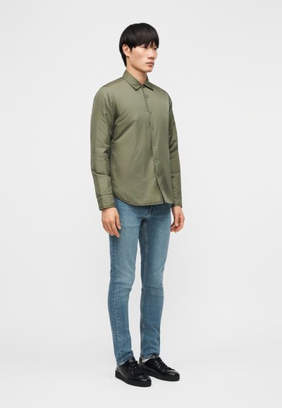 Homme debout portant une veste olive verte boutonnée, un jean skinny bleu et des baskets noires sur un fond clair uni.