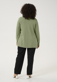 Top verde cu mâneci lungi, cu o croială lejeră, cu tiv bordat, asociat cu pantaloni negri. Modelul poartă sandale albe cu toc bloc.