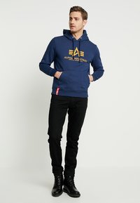 Felpa blu navy con un logo grafico giallo. Presenta una tasca frontale e un cappuccio con cordoncino, abbinata a pantaloni neri e stivali neri.