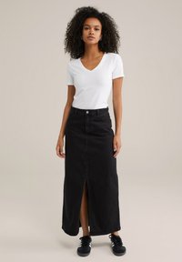 Femme avec des cheveux bouclés portant un t-shirt blanc à col en V, une jupe longue noire taille haute avec fente à l'avant, et des baskets noires avec des rayures blanches.
