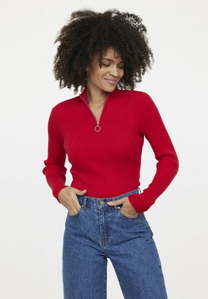 Rode geribbelde gebreide langemouwen top met een halve rits en zilverkleurige accenten, gecombineerd met blauwe high-waisted denim jeans.