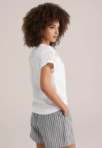 Witte katoenen t-shirt met geborduurde accenten op de schouders, gecombineerd met gestreepte zwart-witte shorts. Model poseert zijwaarts.