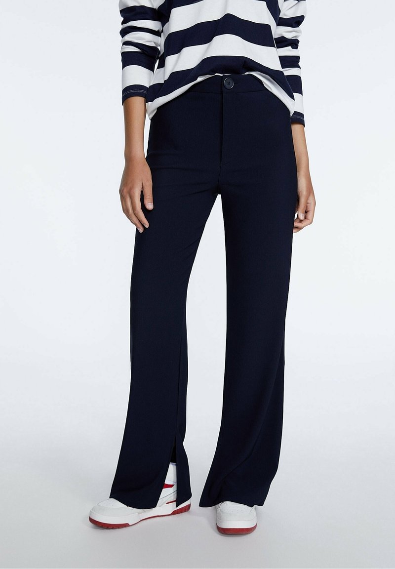 Femme portant un pantalon large bleu marine avec des fentes à l'avant et des baskets blanches, associé à un haut à manches longues rayé bleu marine et blanc.