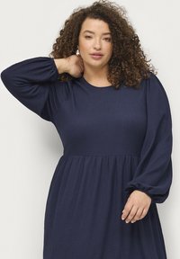 Vestido de manga larga azul marino con tejido texturizado, escote redondo y cintura fruncida. Incluye mangas abullonadas con puños elásticos.