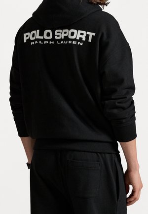 Osoba w czarnej bluzie z kapturem i spodniach, na plecach bluzy biały napis „POLO SPORT RALPH LAUREN”.