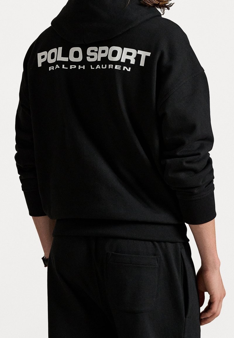 Personne portant un sweat à capuche et un pantalon noirs, avec le texte « POLO SPORT RALPH LAUREN » imprimé en blanc au dos du sweat.