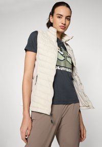 Ľahký puffer vest v krémovej farbe s horizontálnym prešívaním a vysokým golierom; skombinovaný s tmavým tričkom s grafickým dizajnom.