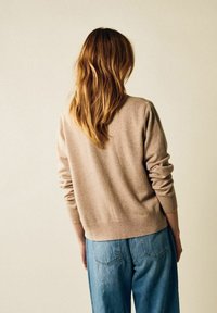 Beige tröja med ribbade muddar och nederkant, kombinerad med ljusblå jeans. Mjuk textur och avslappnad passform synlig från baksidan.