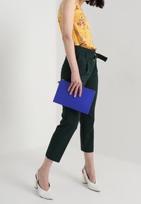 Green tailored pants with a belt, yellow floral top, white heeled shoes, and a blue clutch bag. Smooth textures and structured design.
