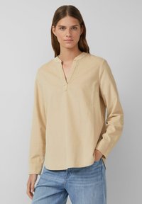Beige Langarmshirt mit V-Ausschnitt, ausgestattet mit einer subtilen Textur und einem abgerundeten Saum. Kombiniert mit blauen Jeans.