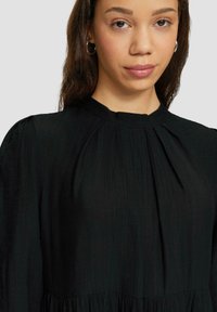 Schwarzes langärmeliges Kleid mit hohem Kragen, gerafften Details und strukturiertem Stoff. Sanfte, fließende Silhouette mit Falten.