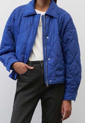 Femme portant une veste matelassée bleu vif par-dessus une chemise blanche et un jean noir, avec une main dans sa poche.