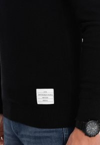 Schwarzer strickpullover mit einem strukturierten Muster. Verfügt über ein weißes Etikett mit Text. Dunkle Jeans sind sichtbar, ergänzt durch eine schwarze Armbanduhr.