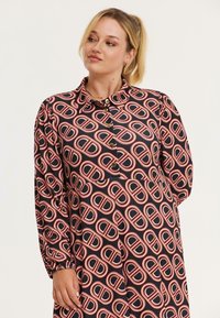 Zwarte jurk met een herhalend geometrisch patroon in rood en wit, met een knoopkraag en puffmouwen, gemaakt van een gladde stof.