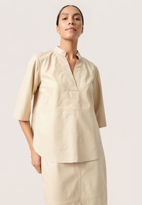 Beige lædertop med kinesisk krave og V-formet udskæring. Har trekvartærmer og en buet kant med synlige sømme.