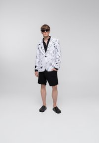 Biały żakiet z czarnym wzorem w krople farby, czarna koszula, czarne szorty i czarne buty bez zapięcia. Model stoi pewnie na tle jednolitego tła.