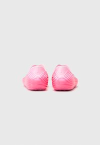 Pink athletic shoes with a wave-like texture on the heel, smooth upper, and recessed logo. Minimalist design with a lightweight, flexible feel.