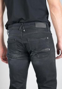 Jean en denim noir avec une coupe slim, un design à cinq poches et une étiquette en cuir sur la ceinture arrière. Décoloration subtile sur le tissu.