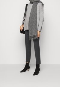 Pull en tricot gris clair, écharpe à carreaux noir et blanc avec franges, pantalon gris foncé ajusté, et bottines noires à bouts pointus.