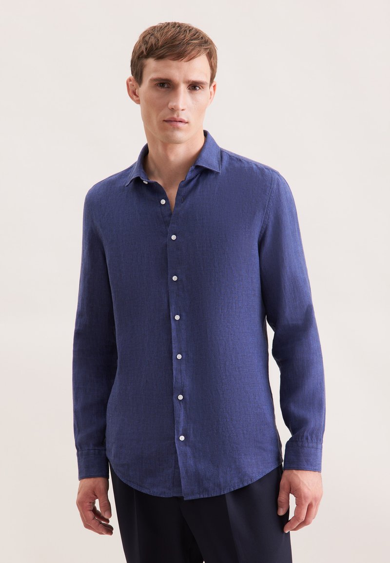 Uomo con camicia blu a maniche lunghe e abbottonata in piedi davanti a uno sfondo chiaro e uniforme.