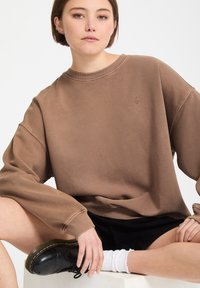 Jeune personne assise en tailleur portant un sweat-shirt marron oversize, un short noir, des chaussettes blanches et des chaussures en cuir noires.