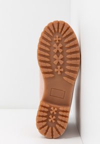 Semelle d'une botte en caoutchouc beige avec des crampons profonds, comprenant des motifs en forme de croix et de barre pour l'adhérence.