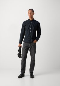 Camisa negra de botones con un brillo sutil, jeans grises y zapatos negros. El sujeto sostiene unos auriculares negros, de pie sobre un fondo liso.