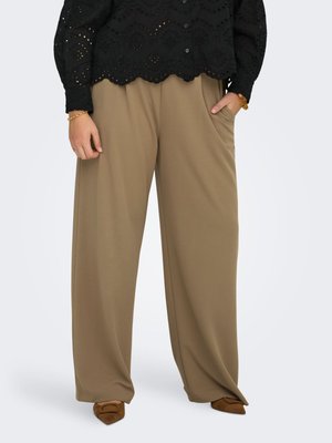 Moteris dėvi plačias smėlio spalvos kelnes, juodą palaidinę su skylėmis, rudos spalvos smailius batus, viena ranka kišenėje, ant riešų nešioja papuošalus.