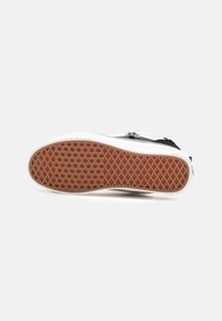 Black sneaker with white rubber sole, featuring a textured brown outsole with a grid pattern, mesh accents, and black laces.