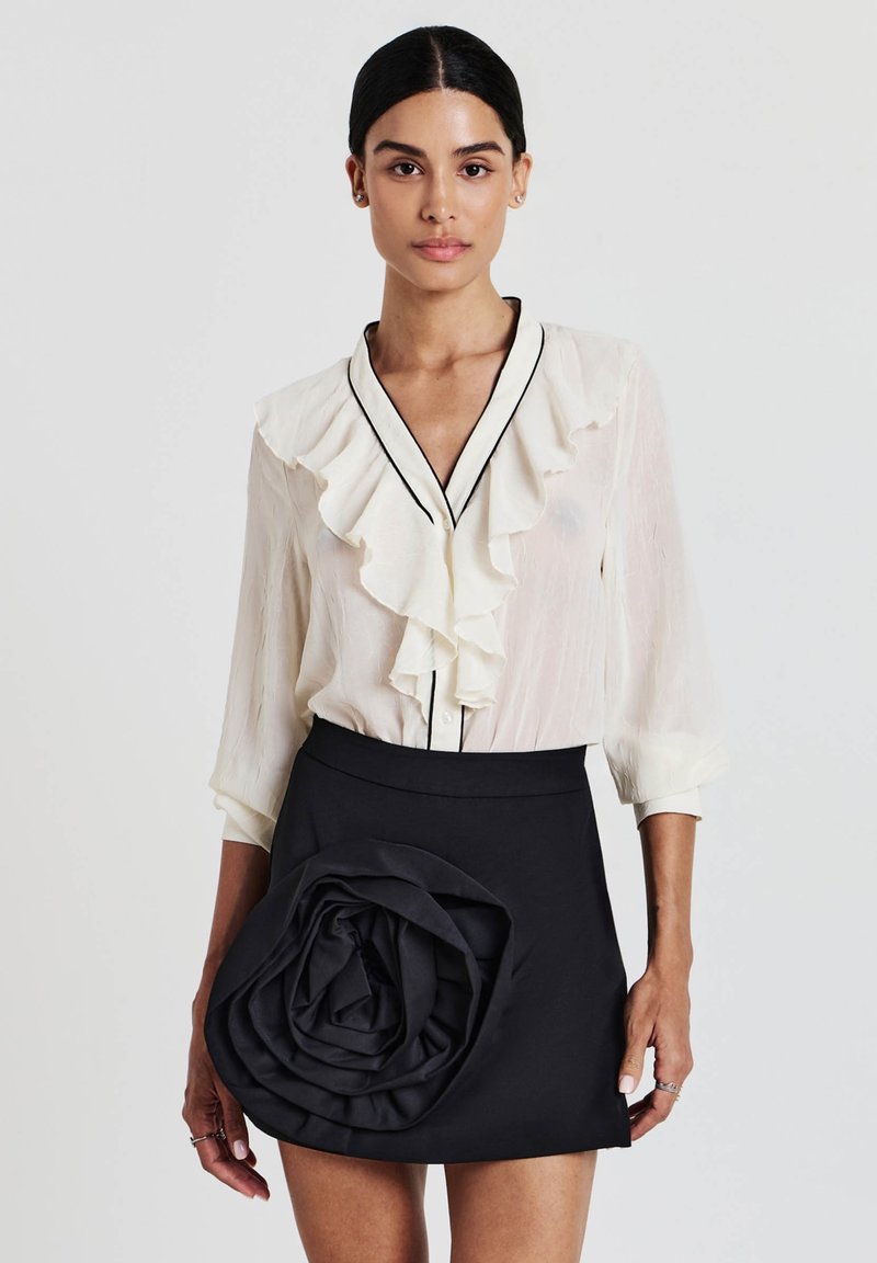 Blusa bianca con colletto a sbuffo e piping nero, abbinata a una gonna nera con una rosa in tessuto scolpita sul davanti.