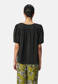 Schwarze Bluse mit kurzen Ärmeln, die eine geraffte Rückendetails und eine glatte Textur aufweist, kombiniert mit gemusterten olivgrünen Hosen, die mit floralen Motiven versehen sind.