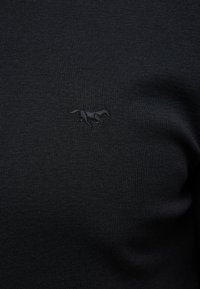 Zbliżenie na czarną tkaninę z subtelnym wyhaftowanym logo biegnącego konia wykonanego czarną nicią na wysokości klatki piersiowej.