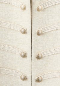 Gros plan sur un tissu texturé crème avec des bordures tressées décoratives et six boutons métalliques ronds disposés en deux rangées verticales.