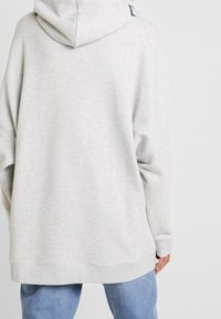 Szara, oversize bluza z kapturem, wykonana z miękkiego materiału. Posiada kaptur, opadające ramiona i ściągacze przy mankietach. Wzór jest gładki, bez widocznych logo.