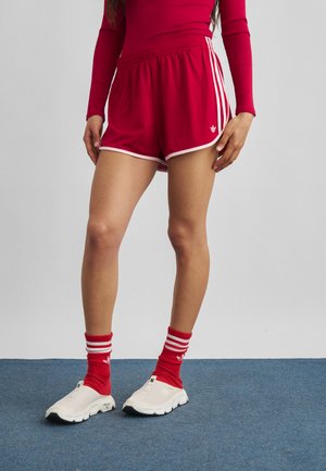 Person iført røde sportsshorts med hvide striber, matchende røde sokker med hvide striber og hvide slip-on sneakers på en blå gulv.