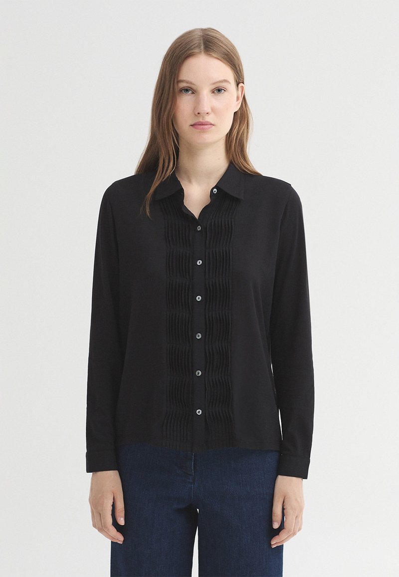 Femme aux cheveux lisses châtain clair porte une chemise noire à manches longues boutonnée avec des plis verticaux et un jean bleu foncé sur un fond uni.