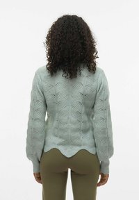 Ljusgrön kabelstickad tröja med scallopdetalj i nederkant och ballongsärmar, som visar strukturerade mönster och en avslappnad passform.
