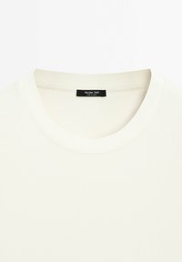 T-shirt couleur crème avec un col rond et une petite étiquette de marque noire cousue à l'intérieur de l'encolure.