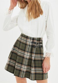 Valkoinen kauluspaita, jossa on pitsikaulus, yhdistettynä vihreään, mustaan ja vaaleanruskeaan ruutuhameeseen, jossa on kaksi mustaa solkea vyötäröllä.