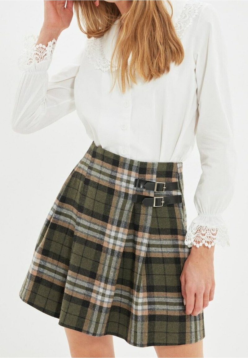 Valkoinen kauluspaita, jossa on pitsikaulus, yhdistettynä vihreään, mustaan ja vaaleanruskeaan ruutuhameeseen, jossa on kaksi mustaa solkea vyötäröllä.