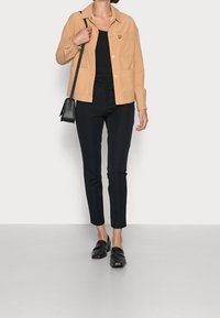 Giacca beige con bottoni e taschino, canotta nera, pantaloni neri su misura e mocassini neri, su uno sfondo neutro.