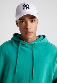Hvid baseballcap med sort "NY" logo, båret med en teal hættetrøje med snor og en afslappet pasform. Glat stoffets tekstur.