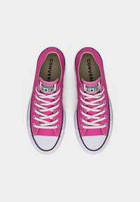 Ζευγάρι ροζ χαμηλά sneakers Converse All Star με λευκά κορδόνια, ελαστικά καλύμματα στα δάχτυλα και μαύρο περίγραμμα, από πάνω σε λευκό φόντο.