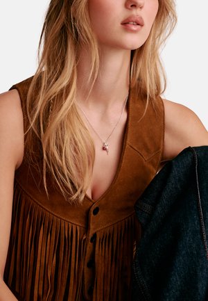 Blonde Frau, die eine braune Wildlederweste mit Fransen und eine silberne Halskette mit rotem Anhänger trägt, hält einen dunklen Denimstoff vor weißem Hintergrund.