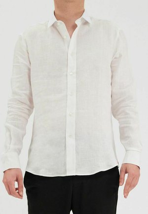 Chemise en lin blanc avec des manches longues, fermeture à boutons devant, et une légère brillance. Présente une texture douce et un ourlet arrondi, associée à un pantalon noir.