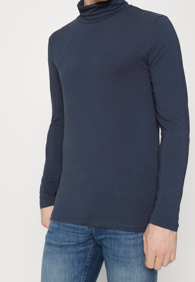 Laineeta, pitkähihaista turtleneck-paitaa pehmeästä, joustavasta kankaasta. Ominaisuuksiin kuuluu istuva muotoilu, sileä pinta ja kääritty kaulus.