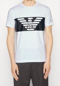 White short-sleeve T-shirt with a large black rectangular logo featuring stylized wings and the letters "GA" on the chest.