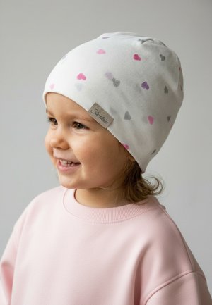 Lächendes Kleinkind mit weißer Mütze mit rosa, lila und grauen Herzmustern und rosa Sweatshirt vor neutralem Hintergrund.