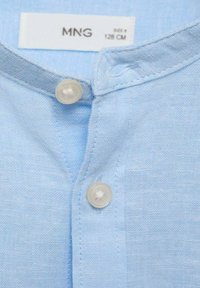 Camicia di lino azzurro chiaro con colletto, caratterizzata da due bottoni bianchi e cuciture a contrasto lungo i bordi. Etichetta con la scritta "MNG, taglia 8, 128 CM."